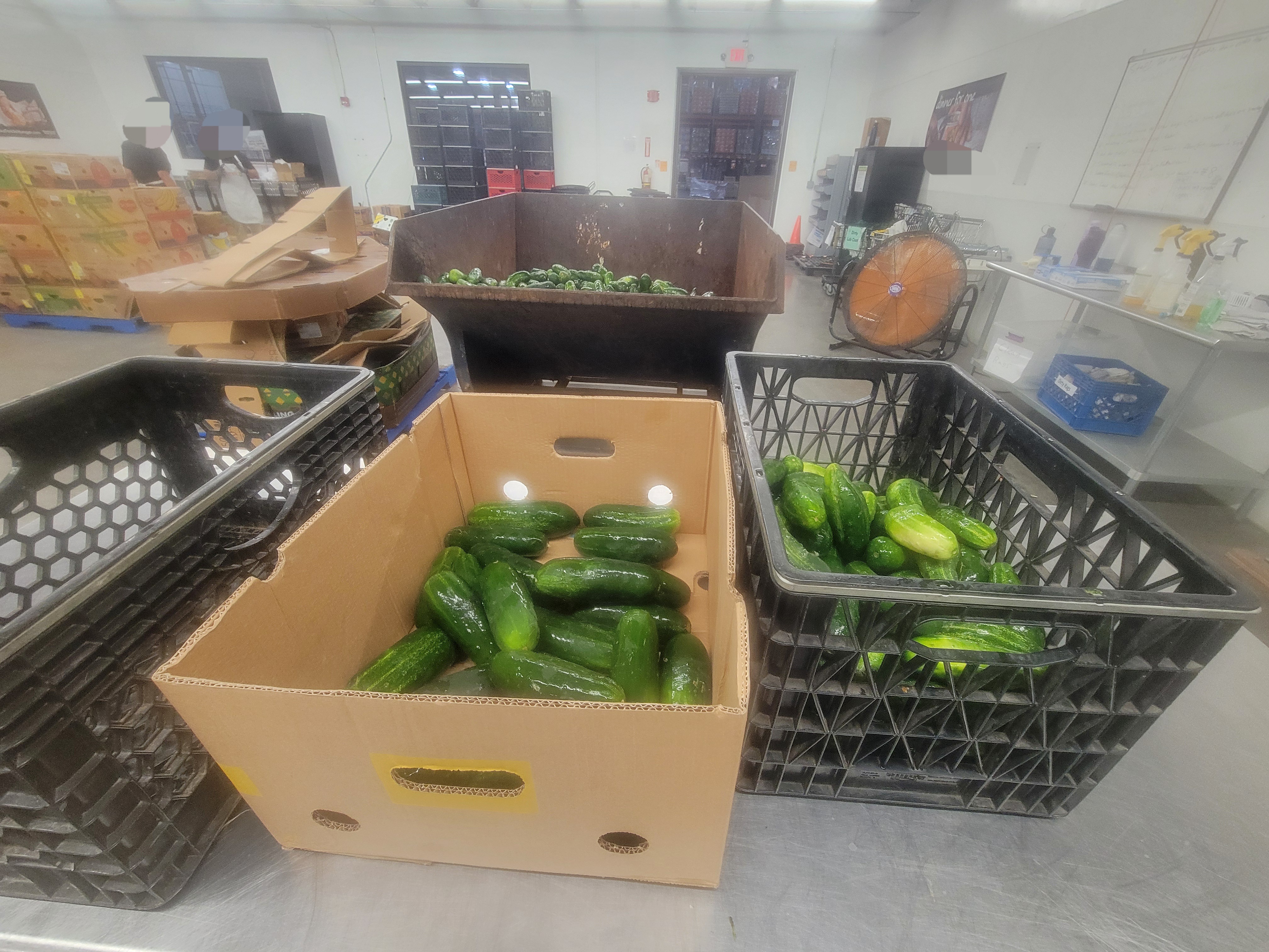 Two crates and one box on metal table with cucumber filled dumpster behind it. One crate and the box has cucumbers in them.